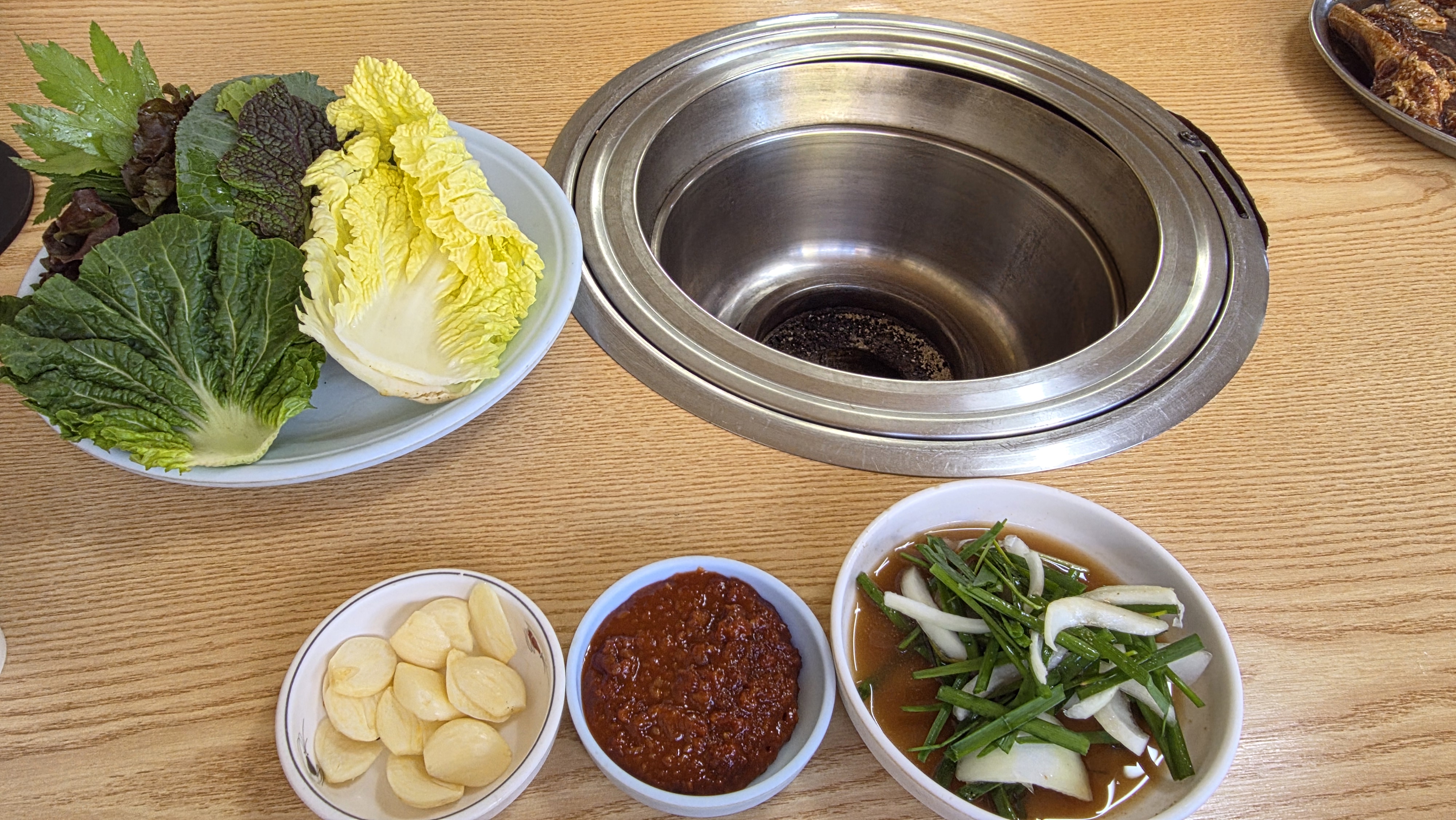 Leafy wraps, cabbage leaves, garlic, red seasoning and chive-onion accompaniments beside the table brazier